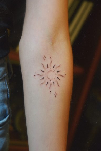 Tatouage de soleil, lune et etoiles ornemental sur l'avant-bras, rayons fins autour d'un cercle central.