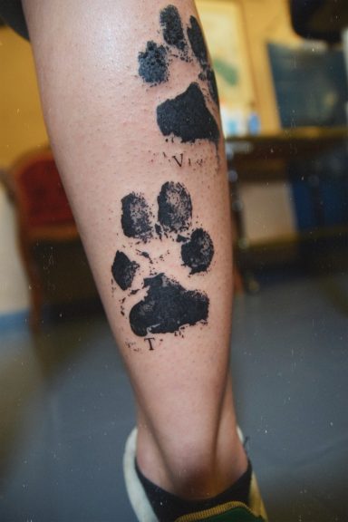 Tatouage de traces de pattes de chiens noires sur le mollet d'une personne.